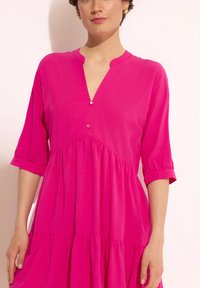 Fuksjowa sukienka z dekoltem w serek i rękawami do łokcia, z dwoma guzikami przy dekolcie oraz warstwowym krojem z miękkiej tkaniny.