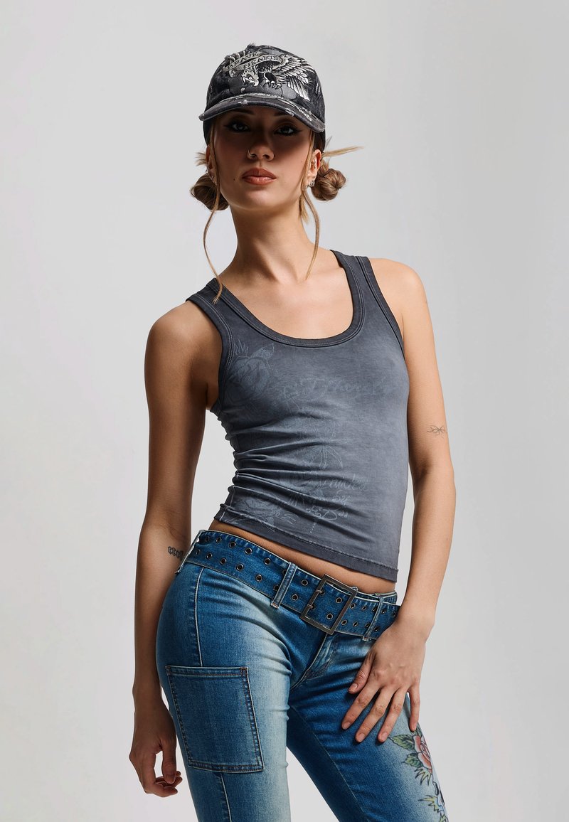 Ung kvinde iført en sort kasket med grafik, ærmeløs falmet sort top, blå jeans med nitter og blomsterlappose, poserer mod en ensfarvet baggrund.
