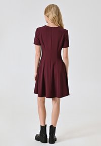 Bordeauxfarbenes kurzärmeliges Kleid mit einem ausgestellten Rock, tailliertem Oberteil und Reißverschluss hinten. Kombiniert mit schwarzen Stiefeletten. Glatte, dehnbare Textur.