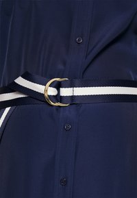 Námořnická modrá košile s hladkou texturou, která má design s páskem a tenkým bílým proužkem a zlatou kulatou sponou. Viditelné detaily knoflíků.