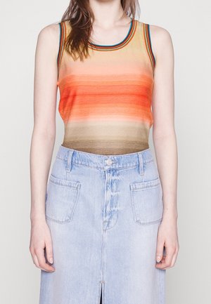 Femme portant un haut sans manches rayé aux tons orange et marron, et une jupe en denim bleu clair avec poches avant et une fente à l’avant.