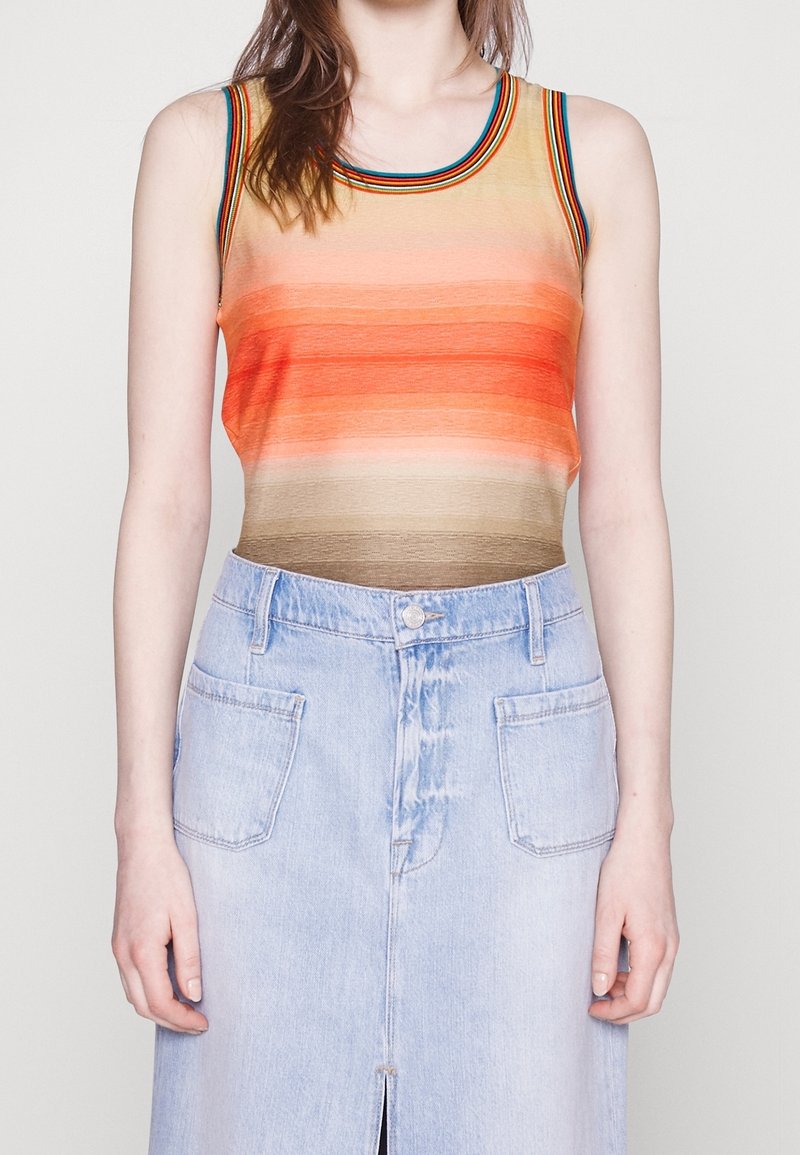 Femme portant un haut sans manches rayé aux tons orange et marron, et une jupe en denim bleu clair avec poches avant et une fente à l’avant.