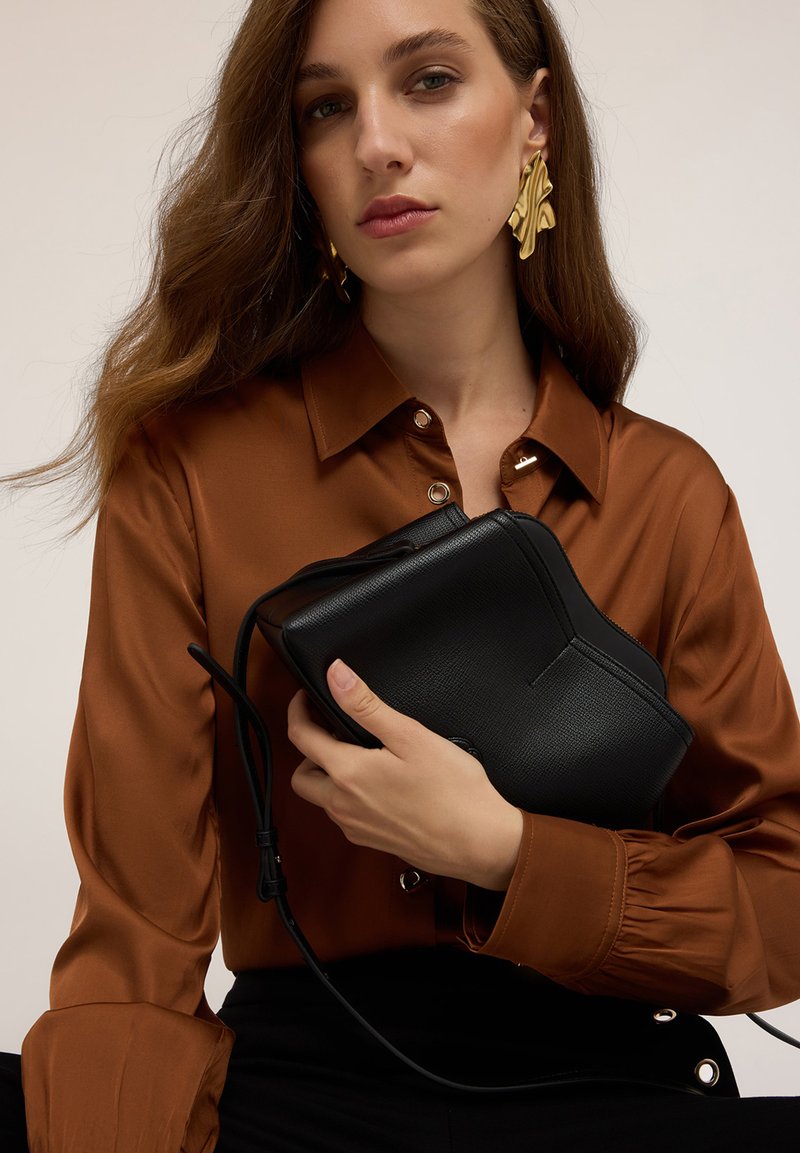 Sort lædertaske med tekstureret overflade, båret på tværs af kroppen og holdt i en hånd. Modellen har en brun skjorte med knapper og en struktureret krave på.