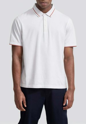 Uomo che indossa una polo bianca a maniche corte con colletto e bordi delle maniche rifiniti in rosso, abbinata a pantaloni scuri, in piedi dritto.