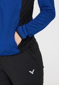 Blå og sort atletisk top med lange ærmer, der har en sidelomme, samt sorte bukser med et gråt logo, der fremhæver lårområdet.
