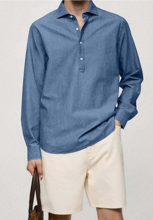 Mężczyzna noszący niebieską koszulę z długim rękawem zapinaną na guziki oraz kremowe szorty, trzymający brązową skórzaną torbę w lewej ręce.