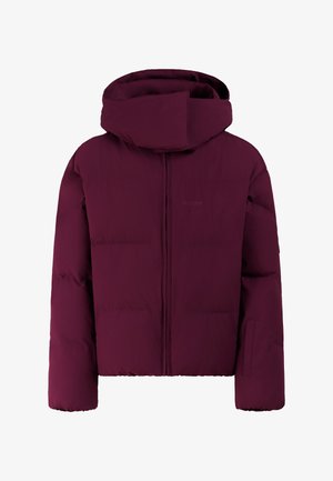 Jonge vrouw staat in een paarse pufferjas, zwarte handschoenen, zwarte broek, gestreepte gebreide muts en roze zonnebril tegen een effen achtergrond.