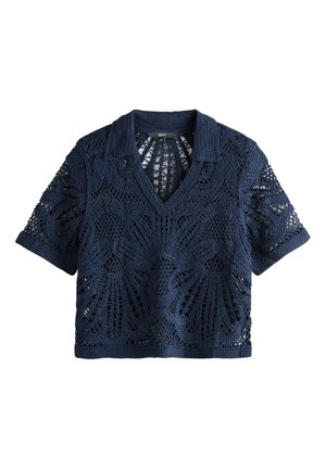 Blonde vrouw die een marineblauwe gehaakte top en losse witte broek draagt, loopt tegen een effen lichte achtergrond.