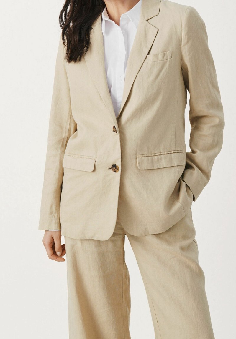 Lichtbeige linnen blazer met twee knopen, een enkele borstzak en meestal revers; gedragen over een wit overhemd met kraag. Gladde textuur.