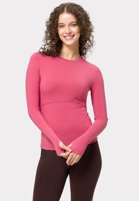 Langärmliges rotes Oberteil aus glattem Stoff, mit taillierter Passform, Daumenlöchern in den Ärmeln und einem runden Ausschnitt. Braune Leggings runden den Look ab.