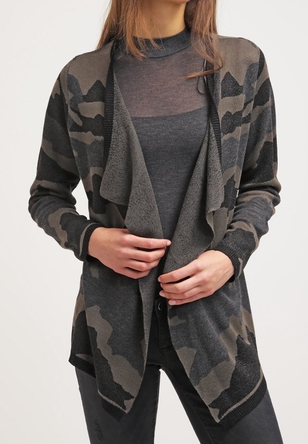 Femme portant un cardigan à ouverture devant camouflage gris foncé par-dessus un col roulé noir transparent, les mains ajustant le devant du cardigan.