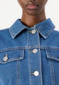 Modra jeans jakna s poudarjenim ovratnikom, srebrnimi gumbi in žepi na prsih s kontrastnim šivanjem. Gladka tekstura, klasičen dizajn.