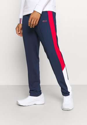 Mand iført navy blå FILA bukser med røde og hvide sidelommer og hvide sneakers, stående mod en ensartet lys baggrund.
