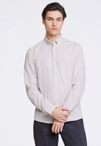Cămașă lungă cu mâneci, de un bej deschis, cu o textură subtilă, având guler și închidere cu nasturi, purtată cu pantaloni închisi la culoare.