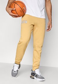 Άνδρας φορά κίτρινες φόρμες με κείμενο "Βοστώνη", μαύρα και λευκά αθλητικά παπούτσια και κρατάει ένα μπάσκετ στο αριστερό του χέρι.