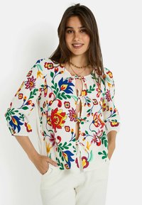 Blusa ligera con estampado floral, de mangas tres cuartos, escote redondo y detalle de lazo en la parte delantera. Los colores incluyen rojo, azul, verde y amarillo.