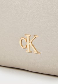 Šviesiai kreminės spalvos tekstūruota odinė rankinė su išsiskiriančiu auksiniu "ck" logotipu. Paviršius yra šiek tiek grublėtas.