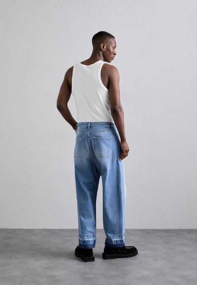 Un débardeur blanc et un jean en denim bleu clair surdimensionné, présentant une coupe légèrement fuselée, avec un ourlet retroussé et des chaussures noires.