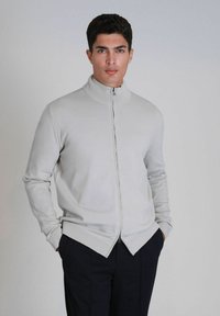Lichtgrijze ritsvest met een hoge kraag, geribbelde details, lange mouwen en een taps toelopende onderkant. Het materiaal lijkt soepel en flexibel.