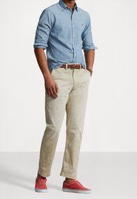 Camicia in chambray azzurro chiaro, pantaloni beige con macchie di vernice, cintura intrecciata marrone e sneakers rosse. Linee pulite e vestibilità casual.