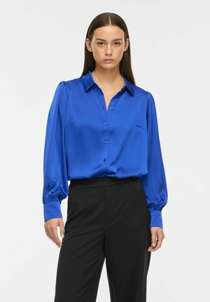 Femme portant un chemisier bleu vif à manches longues boutonné et un pantalon noir taille haute, debout devant un fond blanc uni.