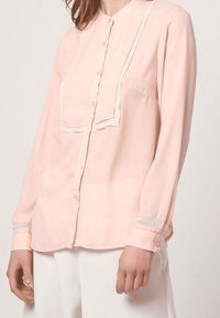 Blusa de manga larga rosa claro con frente de botones, acentos texturizados y un ribete decorativo a lo largo del escote y las mangas.