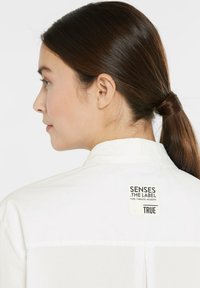 White collared shirt with a tag reading "SENSES .THE LABEL" on the back. The fabric appears smooth with a minimalistic design.