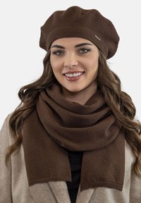 Bérét en tricot marron et écharpe assortie en matériau doux. L'écharpe est drapée autour du cou, mettant en valeur la texture lisse et la constance de la couleur.