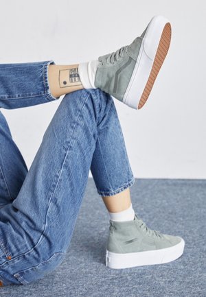 Personne portant un jean bleu clair, des chaussettes blanches et des baskets montantes grises, une jambe croisée sur l’autre, montrant un tatouage rectangulaire à la cheville.
