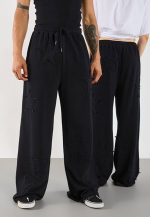 Zwarte wide-leg sweatpants met ster-uitknipingen en versleten randen. Voorzien van een elastische tailleband en trekkoord, gecombineerd met zwarte sneakers.