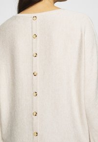 Top in maglina beige con scollo rotondo, schiena aperta e sette bottoni dorati al centro. Tessuto morbido e testurizzato con un motivo sottile.