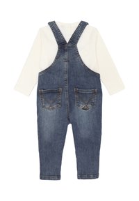 Salopette in denim con bretelle regolabili, di colore azzurro chiaro, due tasche frontali e una maglietta bianca a maniche lunghe sotto.