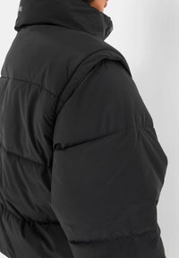 Zwarte pufferjack met hoge kraag en een doorgestikt ontwerp. Heeft grote, gewatteerde schouders en een gladde, glanzende stofstructuur.