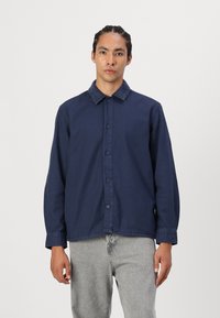 Navy blue long-sleeve shirt with button closure, solid fabric, spread collar, and buttoned cuffs. Worn over light gray trousers.