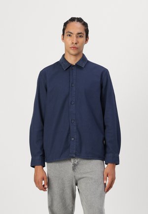 Navy blue long-sleeve shirt with button closure, solid fabric, spread collar, and buttoned cuffs. Worn over light gray trousers.