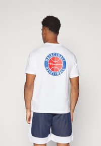 T-shirt en coton blanc avec un graphique de basketball rouge et bleu au dos, accompagné de texte. Tissu léger, col rond, manches courtes.