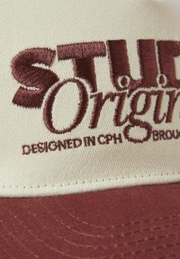 Baseball cap med cremefarvet stof og en maroon skygge. Har broderet tekst "STUD" og "Origin" med sætningen "DESIGNED IN CPH" nedenfor.