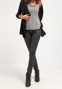 Un cardigan noir porté sur un débardeur à motifs noir et blanc, associé à un pantalon noir ajusté et des bottines. Texture lisse, design ajusté.