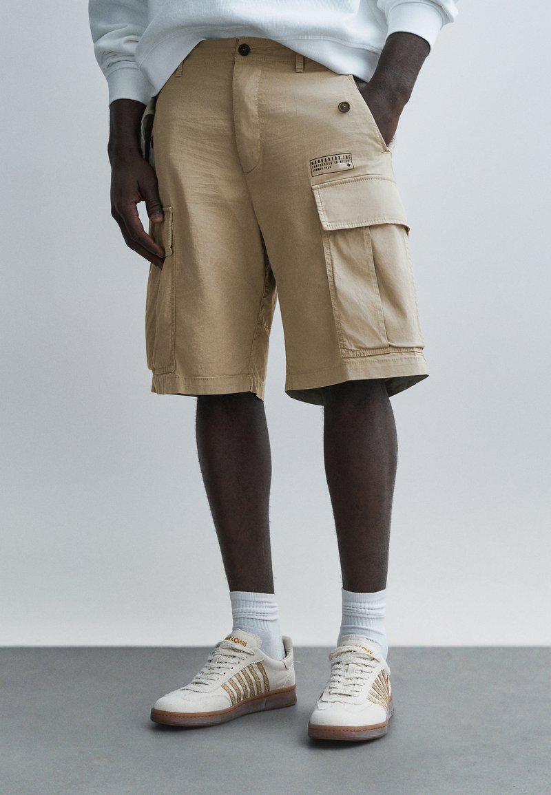 Άνδρας που φοράει μπεζ cargo βερμούδα, λευκές κάλτσ�ες και κρεμ αθλητικά παπούτσια, στέκεται σε γκρι πάτωμα μπροστά σε απλό φωτεινό φόντο.