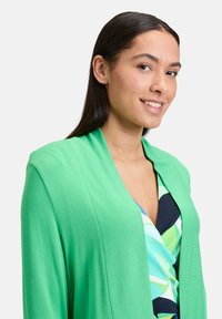 Cardigan vert au design drapé, fabriqué en matériau doux, porté par-dessus une robe multicolore avec des motifs abstraits et un décolleté en V profond.