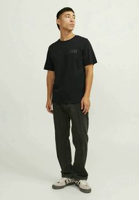 Young man standing, wearing a black Jack & Jones t-shirt, dark jeans, and white sneakers, looking to the side.