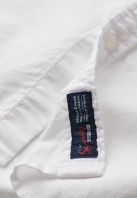 Camisa blanca con una tela texturizada, que presenta una etiqueta azul marino que dice "Superdry" en texto rojo y blanco. Los botones son visibles cerca del cuello.