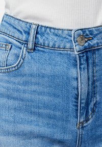 Detailný záber na svetlomodré rifľové rifle v oblasti pásu s gombíkom, pútkami na opasok a predným vreckom, nosené s bielym rebrovaným topom.