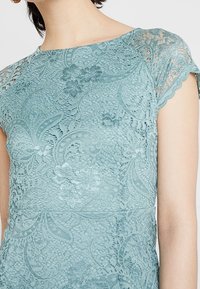 Vestido de encaje en un tono verde claro con mangas cortas con borde en forma de concha, que presenta intrincados patrones florales y detalles texturizados a lo largo de la tela.