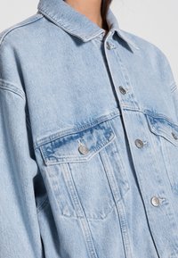 Ljust blå denimjacka med klassisk krage, två framfickor med knappstängning och en avslappnad passform med synliga sömdetaljer.