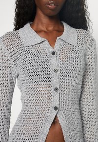Primo piano di una persona che indossa un cardigan in crochet grigio chiaro con bottoni e colletto, che mostra un motivo di lavorazione a maglia testurizzato e la parte centrale del corpo.