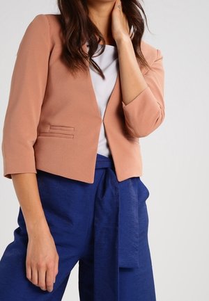 Femme portant un blazer court rose saumon à manches 3/4 sur un haut blanc et un pantalon bleu taille haute avec une ceinture nouée.