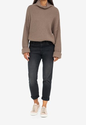 Suéter de punto beige con cuello de cisne, combinado con jeans slim negros. La modelo lleva zapatillas rosa claro, que muestran los puños doblados y un ajuste relajado.