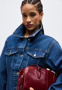 Νέα γυναίκα με πιασμένα μαλλιά φορά χρυσά σκουλαρίκια και ένα μπλε τζιν μπουφάν, κρατώντας μια κόκκινη δερμάτινη τσάντα μπροστά σε ένα απλό φόντο.