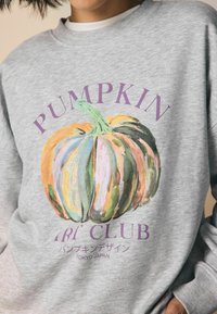 Persona che indossa una felpa grigia con un'illustrazione di una zucca colorata e del testo viola che dice "Pumpkin Art Club" e "Tokyo Giappone" in giapponese.